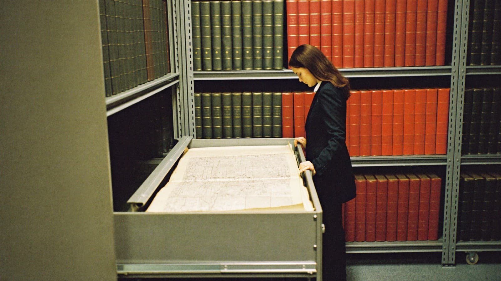 The records vault of a county register of deeds: a woman in a navy suit, seen in full from the side, standing at a wide open flat-file drawer reading a large plat sheet, rolling steel shelves of oversize red and green deed liber volumes behind her.