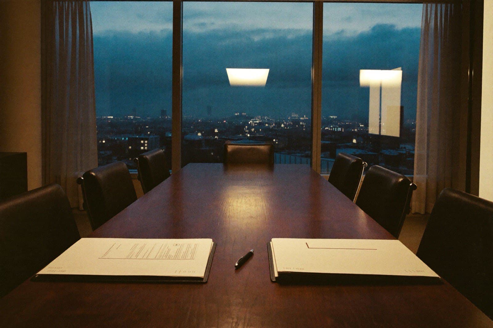 An empty executive boardroom at dusk with two document folders and a pen on the table, city skyline in blue hour beyond the windows.