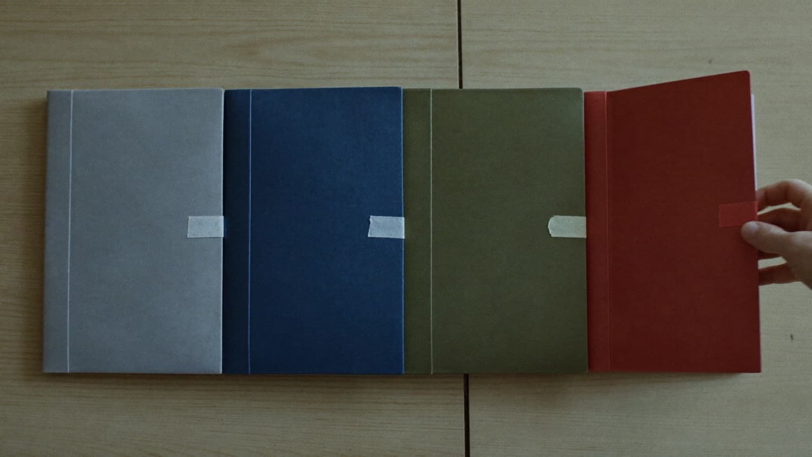 Overhead view of four identical document wallets in four different colours — stone grey, navy, olive and oxblood — laid in a row on a pale oak table, each tied shut with cotton tape, with a hand lifting the fourth clear of the row.