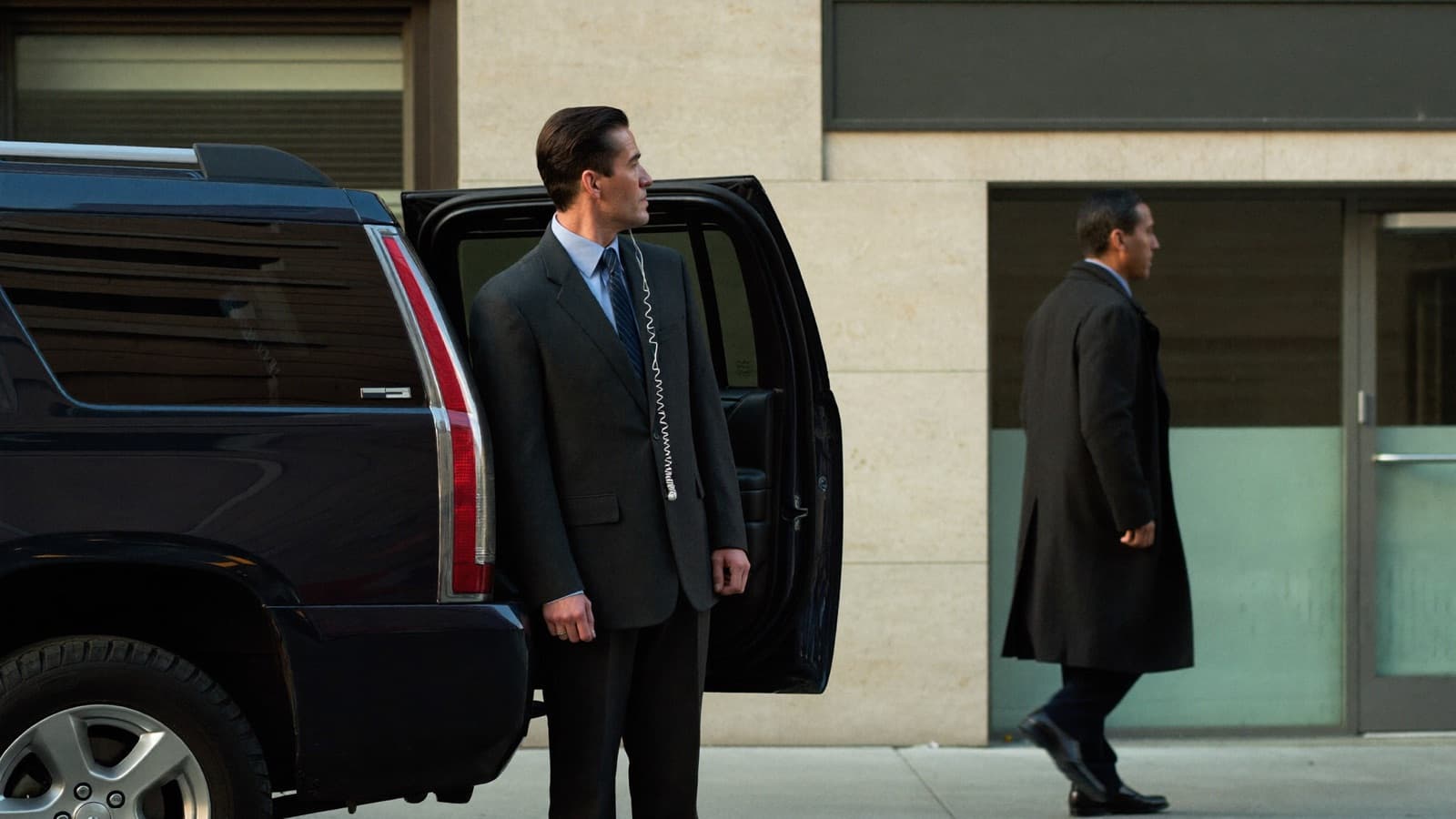 A protective agent in a charcoal suit stands at the open rear door of a dark SUV at a building entrance, head turned in three-quarter view scanning the sidewalk, a translucent coiled earpiece running from his ear behind his lapel, while the principal in a dark overcoat walks from the vehicle toward the entrance doors.