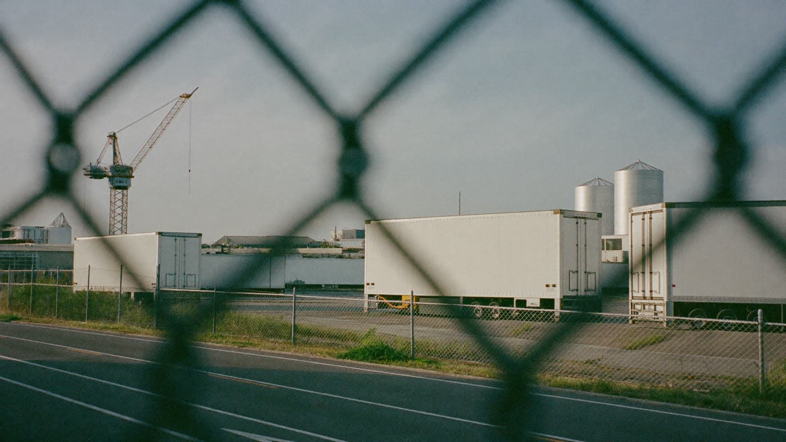 A competitor's distribution yard observed from a public roadside through a chain-link perimeter fence: unmarked white trailers standing at loading bays, a conspicuously empty stretch of yard, and storage silos and a lattice crane beyond.