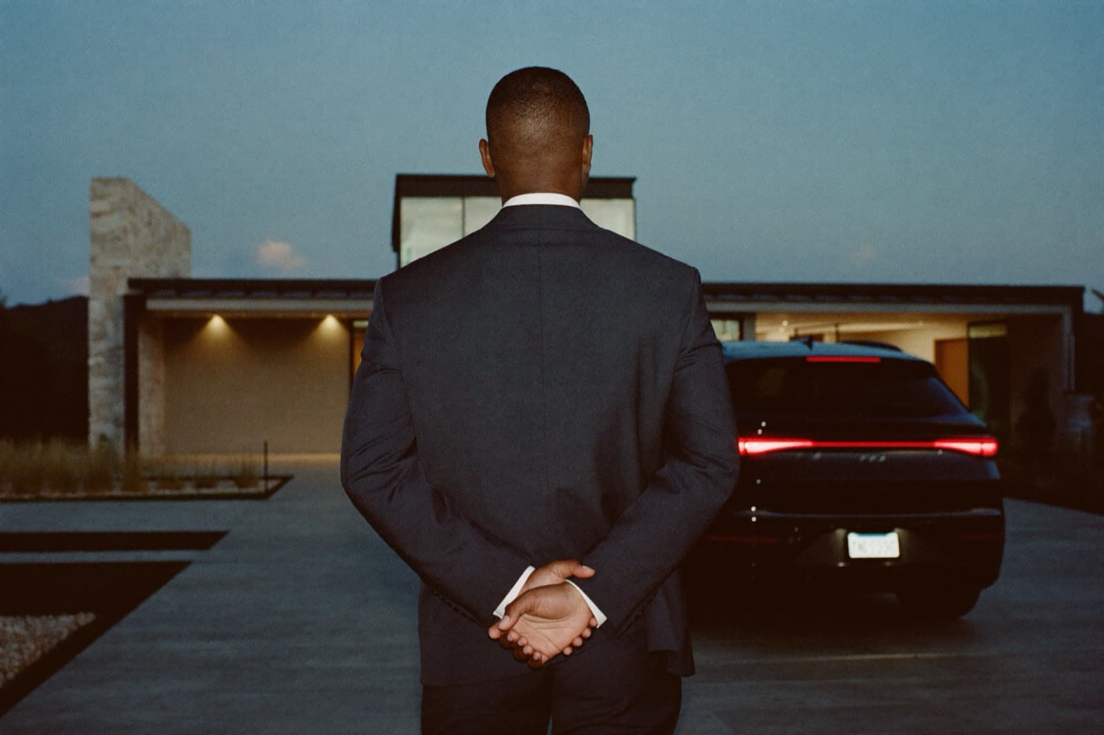 A close-protection professional in a tailored suit, seen from behind at dusk, standing watch over the courtyard of a modern private residence as a black SUV waits on the driveway.