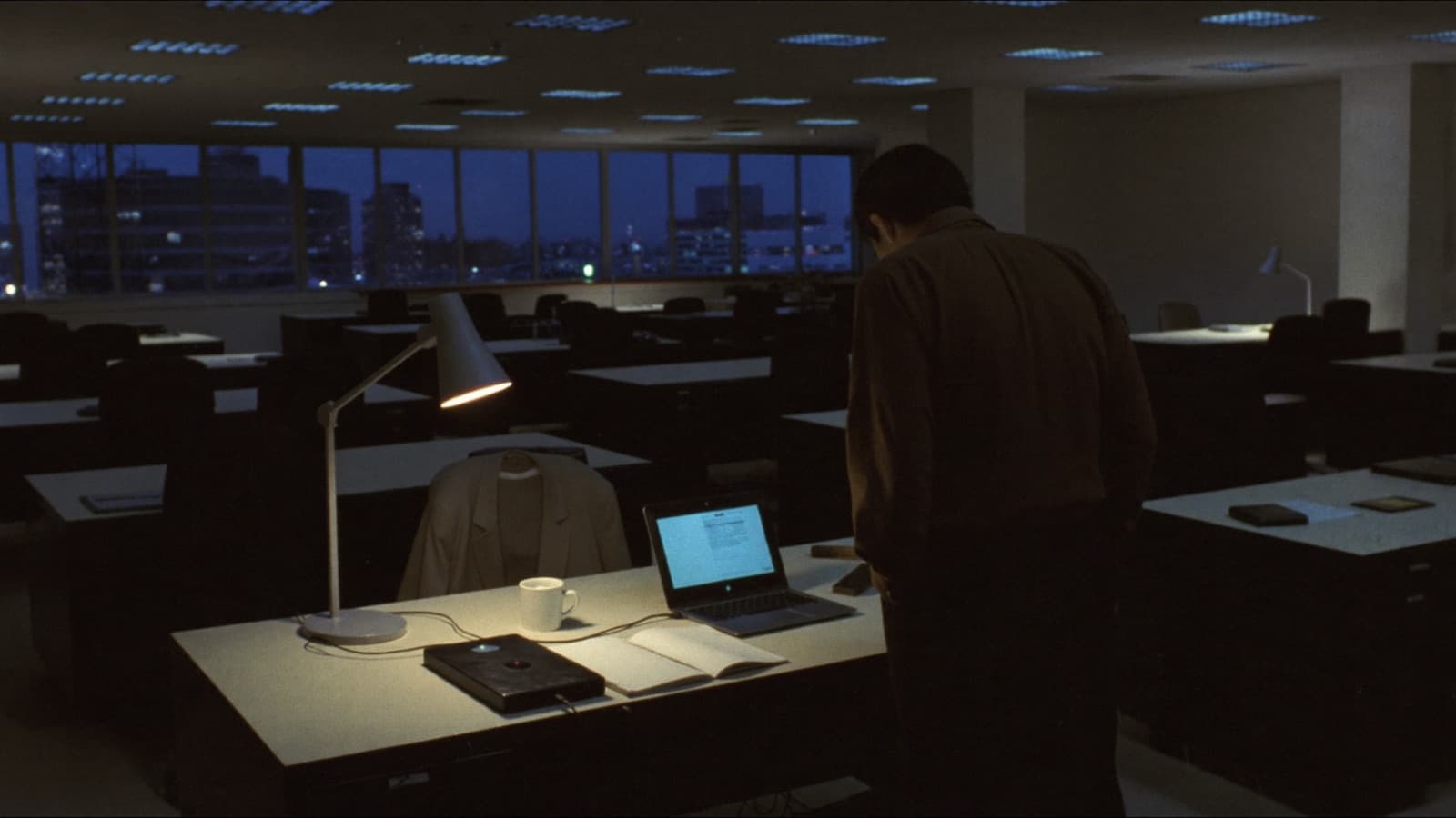 An empty open-plan office at night with every ceiling light off. One desk is lit by a single lamp: an open laptop is tethered by cable to a plain external drive, beside a coffee mug, an open notepad and a jacket still hanging over the chair. A technician stands over the desk with his back to the camera, waiting while the copy runs.