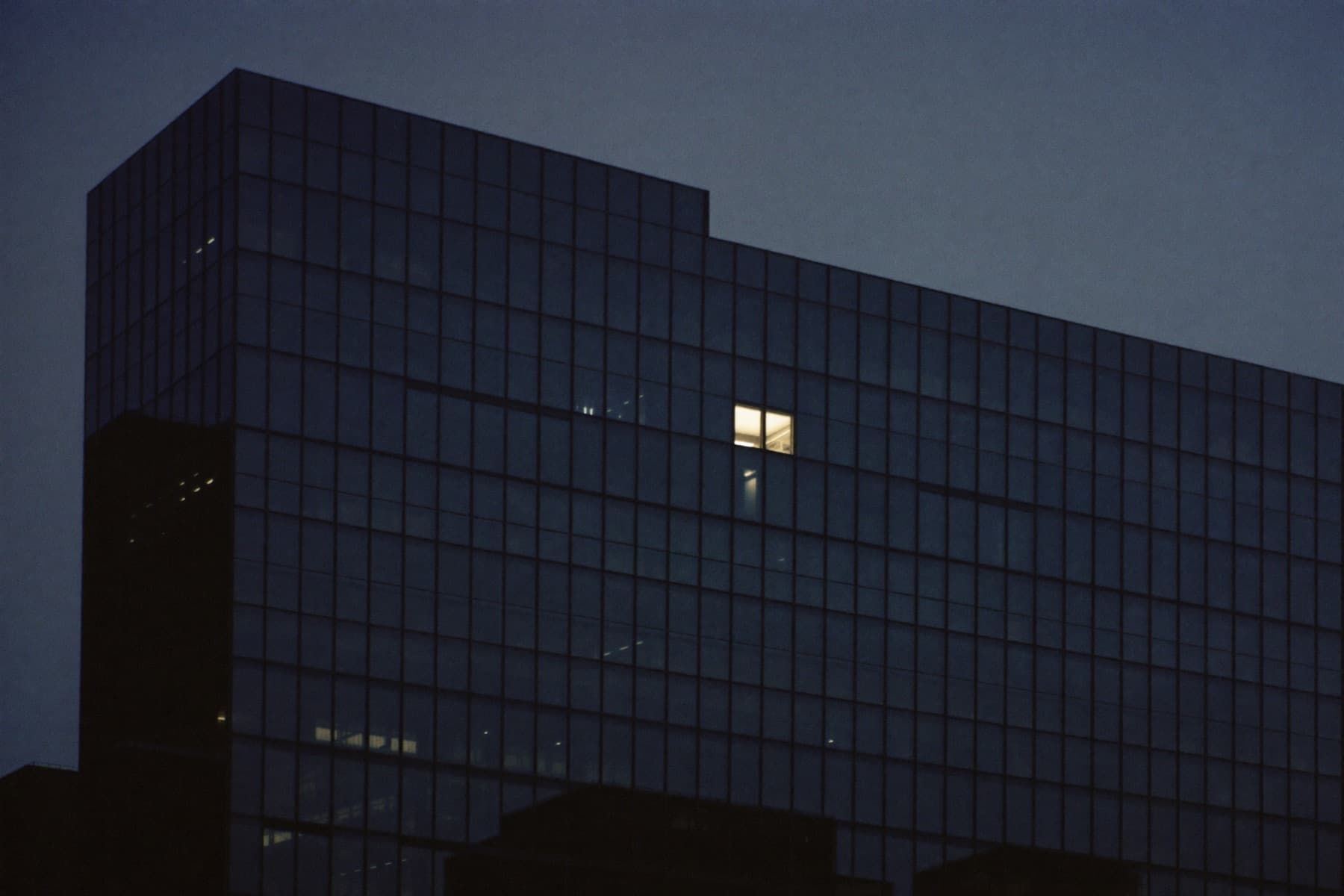Dark glass facade of a modern office tower at dusk with a single office window lit on an upper floor.