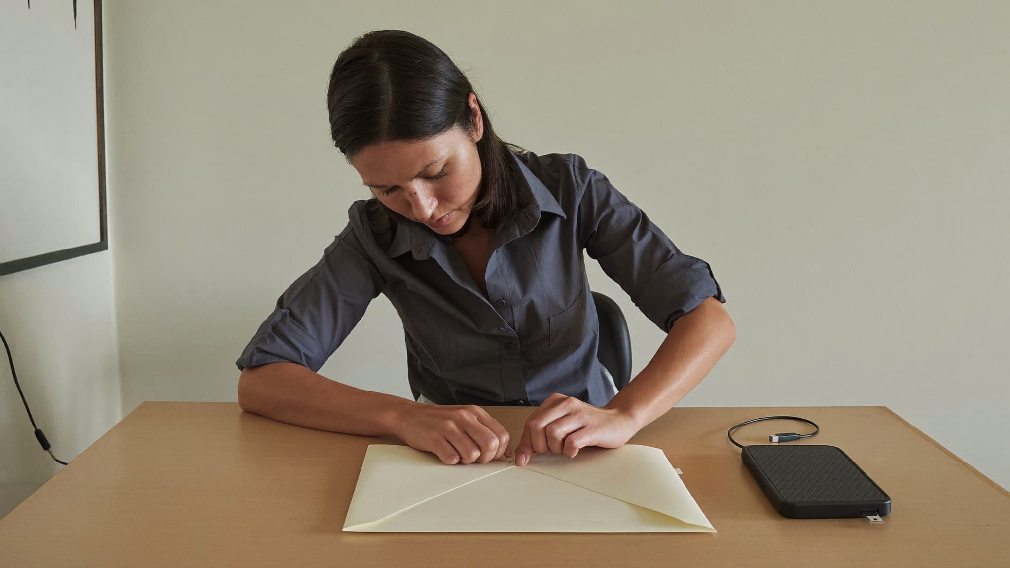 An investigator at a bare desk in a plain workroom, seen three-quarter with her face visible and her eyes on her hands, pressing the flap of an unmarked manila envelope closed, the small external hard drive it will hold lying on the desk beside her with its cable coiled next to it.