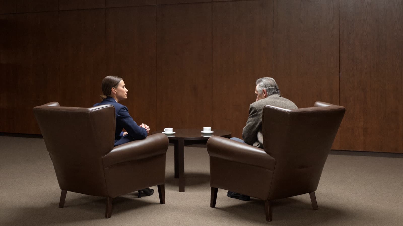 A quiet corner of a private club library with bare wood-panelled walls: a woman investigator in a navy suit and a grey-haired man in a tweed jacket, a former colleague of the executive under review, sit facing each other in two leather armchairs across a low table with two coffee cups, both seen in full, in conversation.