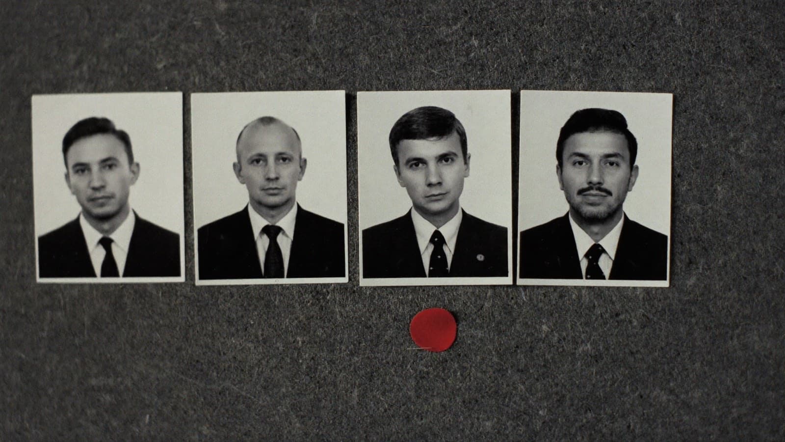 Four passport-format black-and-white portrait photographs of four different middle-aged men in dark suits, pinned in an evenly spaced row on a dark grey felt board, with a single small red adhesive dot marking the board beneath one of the four.