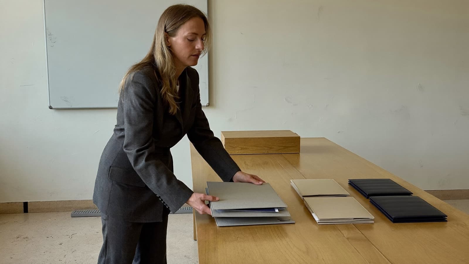 A woman in a charcoal suit, seen in full from the side in a bare daylit meeting room, lowering a closed grey document wallet onto a long oak table to complete the third of three separate groups of files already laid out along it.