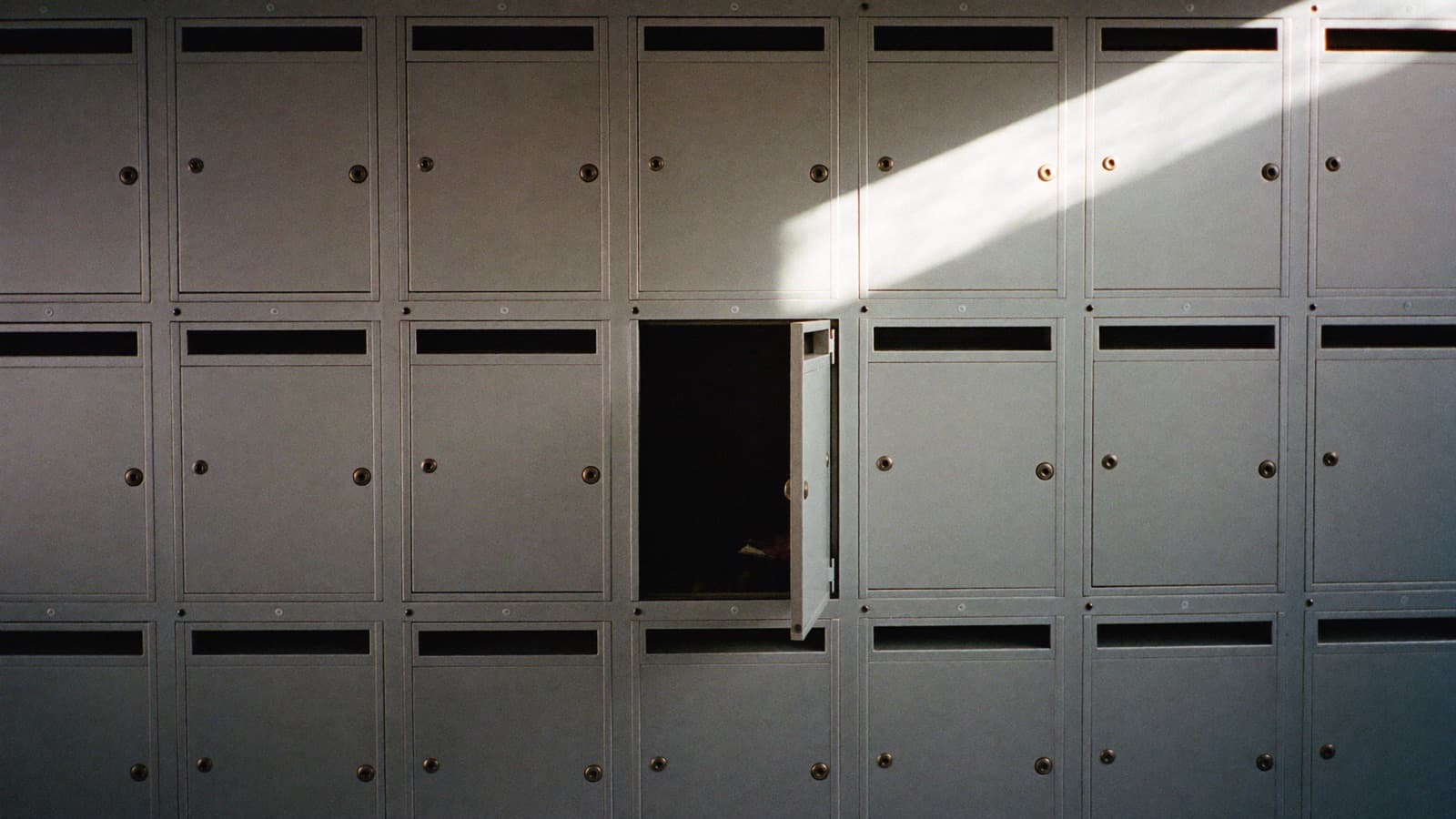 A wall of small unmarked private mail-drop boxes in a mail-forwarding storefront, raked by a diagonal shaft of daylight, one compartment standing open on a completely empty dark interior.