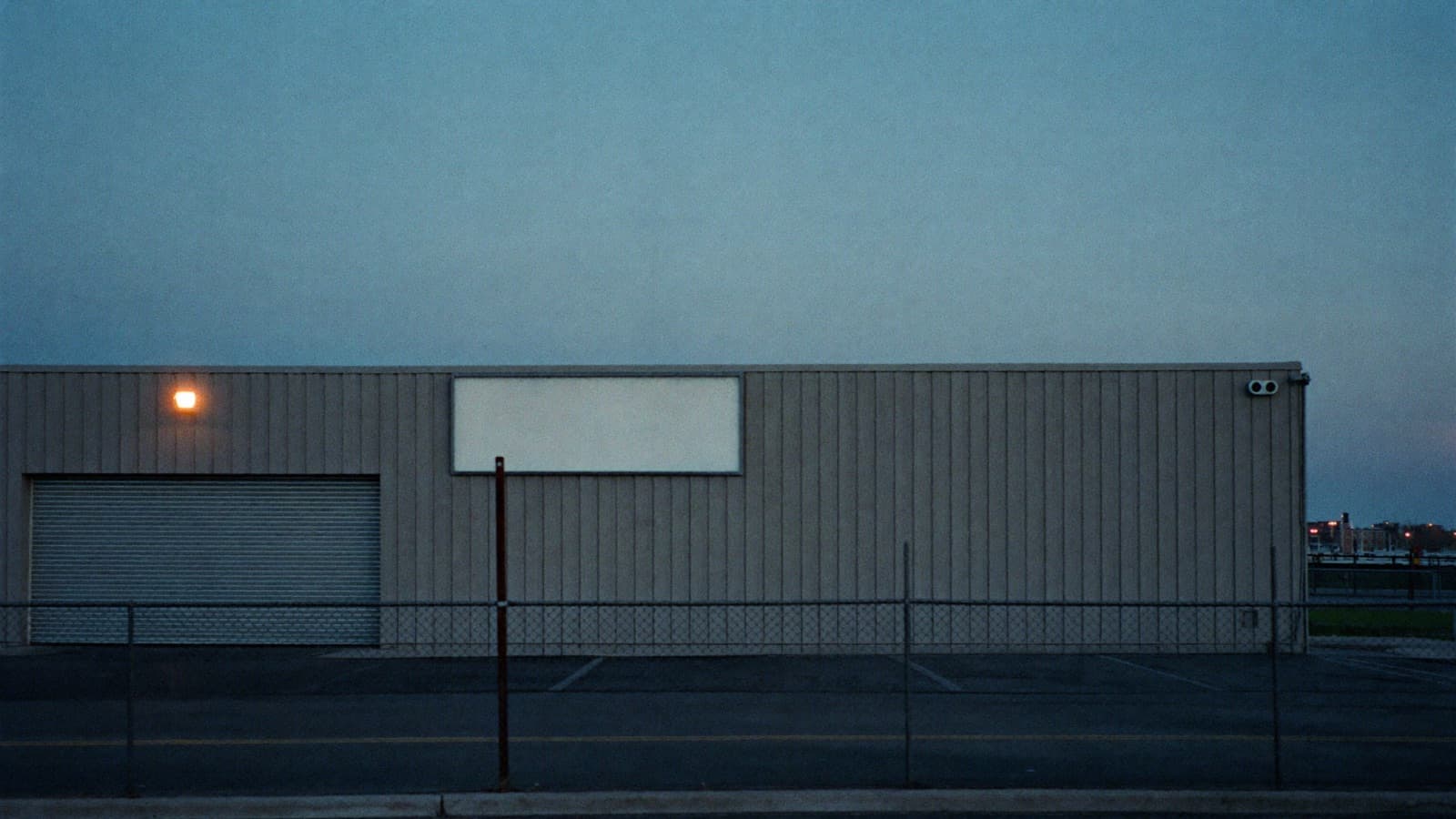 A small industrial supplier warehouse at blue hour seen from behind a chain-link fence: corrugated cladding, a closed roller shutter, an empty parking lot and a blank unbranded signboard by the gate.